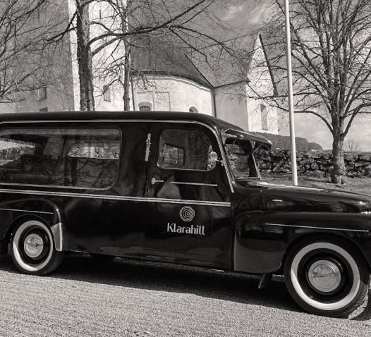 Svartvit veteranbil med texten "Klarahill" parkerad framför en kyrka och träd utan löv.